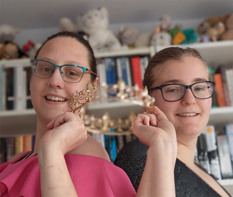The Importance of Neurodiversity Representation in Fiction - Beth and Sifa are two white women standing back to back in a room witha white bookcase filled with books behind them. They are holding a gold crown up in the air between them. They both have dark hair pulled back from their faces and both wear glasses. Beth is on the left and is wearing a pink t-shirt and teal rimmed glasses. Sifa is a few inches shorter than her friend and is wearing a black t-shirt and black rimmed glasses. They are both smiling at the camera.