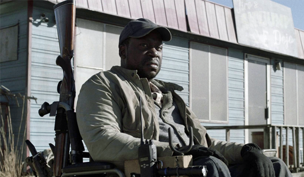 It's time to talk about Disabled actors - A screencap of Daryl Mitchell as Wendell in Fear the Walking Dead Season 5. He is a black man sitting in a wheelchair in front of an outdoor building. He is wearing grey clothing and a dark baseball cap. He has a grim expression on his face and is staring into the distance off camera. There is a shotgun attatched to the back of his wheelchair.