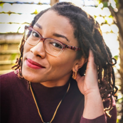 About the author: Tracy Deonn - A black woman sitting outside. She has brown eyes and her dark hair is styled into braids. Her head is to the right and her right hand is lifted to touch her hair. She is wearing a dark red long-sleeved top, glasses, gold hoops and a gold chain pendant.