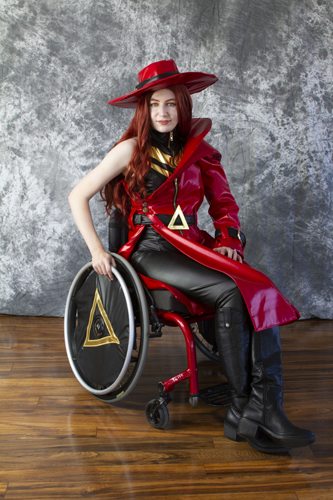 #GeekDis Interview with Colleen Cosplay! - Colleen as Carmen Sandiego. She has auburn hair and is wearing a red wide brimmed hat. She is wearing a red leather coat with a black belt and a hold triangle buckle. Her coat is half off one shoulder, revealing a black sleeveless top with a gold zigzag design. She is wearing black leather pants and black leather boots. She is sitting in her wheelchair and the wheel of her wheelchair has a black cover with a gold triangle on it. She is facing the camera and smiling.