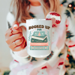 Booked Up For The Holidays Christmas Mug - A white female model holds out a white mug with a teal and pink design of a pile of books. Stars and leaves decorate the area around the books. Above the books is text that reads "Booked up for" and text on the book spines continues the sentence "for the holidays".