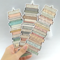 Bookmark Book Tracker - A hand holds out three bookmarks to display them. The bookmarks are printed with a pile of books design and the edges are cut in the shape of books. The books on each bookmark are various colours, and the spines of the first two bookmarks are blank. The spines on the third bookmark has handwritten titles to show how to use the bookmark as a book tracker. A white tassel is connected to the top of each bookmark.