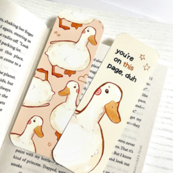 You're on this Page Duck Bookmark - The front and back view of a bookmark displayed side by side on a book. The side on the left shows a design with of white ducks on a peach background. The other bookmark shows a white duck on a cream background with text above it that reads "you're on this page, duh" in black. The word "this" is highlighted in orange. There are two stars in the top right corner.
