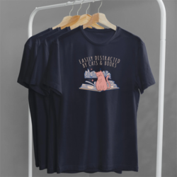 Easily Distracted by cats and books T-shirt - A rack of navy blue t-shirts. The facing t-shirt shows a design of a cat sitting among books printed in pink and blue. Above the design is text that reads "Easily Distracted by cats & books".