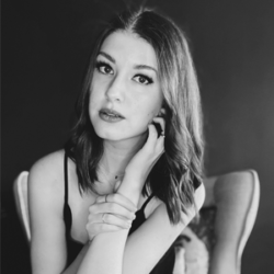 About the author: Kelly Andrew - A black and white photo of a white woman with dark shoulder length hair styled with a side parting. She is sitting in a chair in front of a dark wall, and is leaning forward with her arms resting on her knees. Her right arm reaches up to her face, her fingers resting against her cheek, with her left hand holding onto her right wrist. She is wearing a black top with thin straps and is looking directly at the camera.