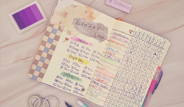 March 2024 Wrap-Up - Flower Pot spread in my Final Fantasy XIV bullet journal - A bullet journal is laid open on a pale pink marble background. The journal shows a spread for flower pots in Final Fantasy XIV, with paper used to decorate the page and ink stamps used to print the title 'flower pots'. Over a spread of two pages are handwritten details about mounts from the video game. Around the journal are various stationery items. To the left, is a small rectangular ink pad with four shades of purple ink. Above the journal, a small purple rectangular post-it note pad has a handwritten message that reads '#jgbbujo' in dark blue in. On the right side of the journal, there are two gel pens, one pink and one dark blue. Below the journal, there is a trio of washi tapes piled together and a teal and white fountain pen unlidded. The lid is sitting next to it.
