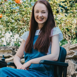 About the author: Pippa Stacey - A young white woman with long straight brown hair. She is sitting in a black wheelchair and is wearing a white short-sleeve top beneath a pair of denim dungarees. Her arms are resting on the armrests of her wheelchair, and her hands are placed together in her lap. She is sitting in a garden and there are plants behind her. She is smiling widely at the camera, and the image is cropped at knee height.