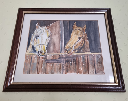 A framed watercolour painting hanging on a pale yellow wall. The painting is of two horses standing side by side in a partitioned stable stall with their heads hanging out over the doors. On the left is a pale grey horse that is facing forward, and on the right is a bay horse, its head turned to look at its neighbo