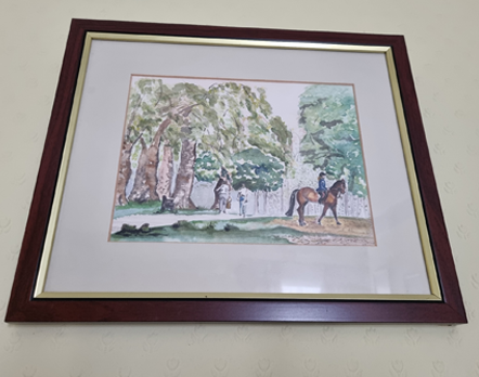 A framed watercolour painting hanging on a pale yellow wall. The painting is of a scene in a park showing a white footpath framed by trees on the left side. A black iron bench sits part way along the path, and in the distance a couple is walking away. In the foreground, there is grass and a dirt path. On the dirt path beneath a tree a female rider with long dark brown hair, riding hat and blue jacked is riding a chestnut horse. They are facing right, away from the couple.