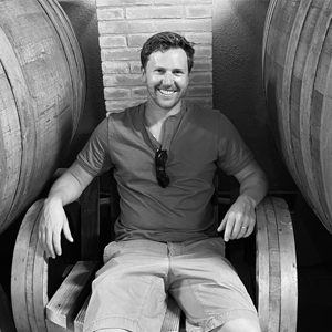 About the author: D.H. Hoskins - A greyscale photo of a white skinned man sitting in a chair made of large barrels, with a barrel on each side of the chair. He has short dark hair, is unshaven and is wearing a dark-coloured long-sleeved top and light-coloured shorts. A pair of sunglasses hang from the neckline of his top. He is smiling at the camera and sitting in a relaxed position. 
