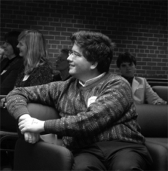 About the author: James Bow - A candid black and white photo of a white man with dark hair. He is sitting watching something off to the side, his head and upper body turned the right. He is wearing glasses. a striped sweater and dark pants.