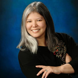 About the author: Julie Kagawa. - An asian woman standing in front of a blue background. She has shoulder length hair; the left side is grey and the other side is dark. She us wearing a long-sleeve black top and has her arms crossed over her chest. She is looking directly at the camera and smiling. The image is cropped to show her head and upper body.