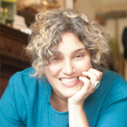 About the author:, Maia Toll - A white woman with dark curly hair parted in the middle. She is sitting indoors and is smiling at the camera. Her right hand is propped up underneath her chin. She is wearing a bright teal top.