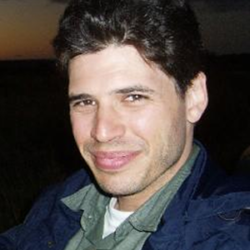 About the author: Max Brooks - A selfie of a white man with short dark hair. He is wearing a dark jacket over a grey collared shirt with a white t-shirt underneath. He is looking directly at the camera with his head turned slightly to the left, and is smiling. The image is cropped to show head and shoulders.